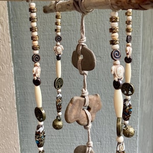 Puede incluir: Un móvil de viento artesanal con una rama de madera natural con hilos colgantes de cuentas y elementos de piedra. Las cuentas son de color crema, marrón y dorado. El móvil de viento incluye pequeñas campanas y formas de piedra.