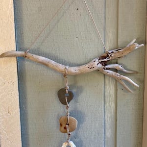 May include: Driftwood wall hanging featuring a natural branch with a light brown hue. Three elements are suspended from the branch: two stones and a seashell. The stones are gray and tan, and the seashell is white.