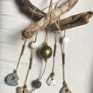 May include: Driftwood wall hanging with natural elements. Two pieces of driftwood are tied together with twine. Hanging below are shells, stones, a brass ball, and a feather, all suspended by twine and brass accents.
