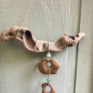 Puede incluir: Móvil de madera flotante con una rama natural y desgastada. Dos piedras marrones talladas cuelgan de la rama con una cuerda, acentuadas con pequeñas cuentas verde claro. El móvil cuelga de una fina cuerda blanquecina.