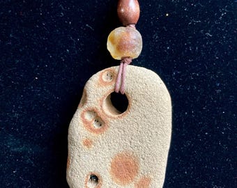 May include: A handcrafted necklace featuring a natural stone pendant with a large central hole and smaller perforations. The pendant is beige with brown spots and is suspended from a brown cord with wooden beads. The necklace is displayed against a dark background.