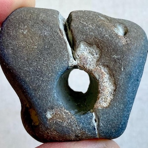 Könnte beinhalten: Eine weiße Keramik-Tasse mit schwarzem Henkel und dem Text "But First, Coffee" in fetter, schwarzer Schrift. Die Tasse steht vor einem verschwommenen Hintergrund, der eine gemütliche Umgebung suggeriert.