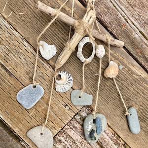 Puede incluir: Móvil artesanal de madera flotante con diversas conchas marinas y piedras grises suspendidas con hilo. La madera flotante está dispuesta en forma de cruz, con las conchas y piedras colgando a diferentes longitudes. Se muestra sobre una superficie de madera envejecida.