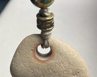 May include: A handcrafted pendant featuring a smooth, light-colored stone with a central hole, strung on a beige cord. The pendant is accented with a blue and brown ceramic bead and a brass-colored metal bead. The stone has a natural, organic shape.
