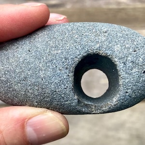 May include: A smooth, oval-shaped gray stone featuring a single, circular hole drilled through its center. The rock has a speckled, textured surface with a natural appearance, showing a consistent dark gray color throughout the entire object.