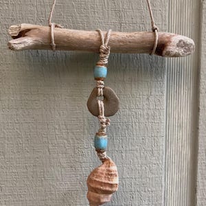Puede incluir: Móvil artesanal de madera flotante con una rama de madera natural, cuerda beige, cuentas turquesas, un anillo de piedra y una concha. El móvil está suspendido por una cuerda beige.