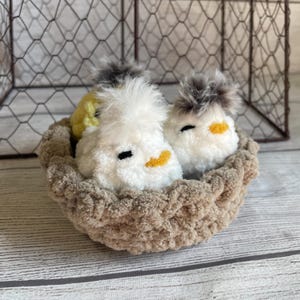 May include: Three fluffy yarn chicks in a brown yarn nest. The chicks have white and yellow yarn bodies with black eyes and orange beaks. The nest is made of brown yarn and has a woven texture.