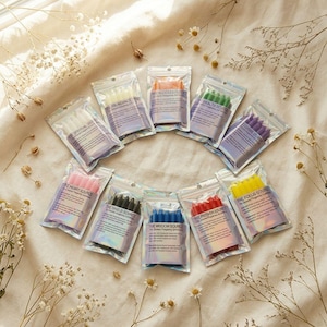 May include: Ten small, clear, resealable pouches arranged in a circle, each containing a set of colored wax candles. The labels on the pouches feature text including titles like The Abundance Source, The Focus Source, and The Shield Source.