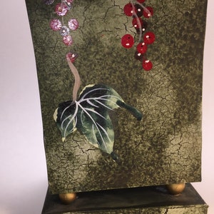 May include: A green ceramic vase with a distressed finish and painted red berries and green leaves. The vase has a square base with gold accents.