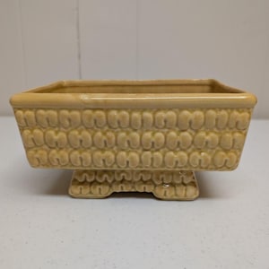 May include: A rectangular ceramic planter with a glossy light yellow finish. The exterior features a repeating embossed pattern of small, rounded, U-shaped links arranged in horizontal rows. The planter sits on a raised pedestal base.