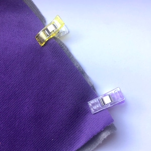May include: Close-up of purple fabric with a grey edge, held together by three clear plastic sewing clips. One clip is yellow, and two are purple. The fabric is a solid colour and appears to be a knit or woven material.
