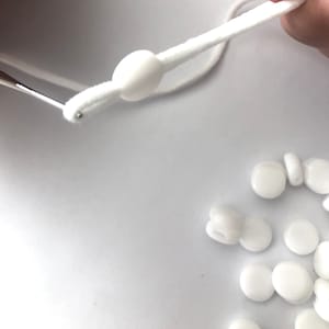 Może przedstawiać: Białe plastikowe stopery do sznurków do masek. Stopery są okrągłe i mają otwór w środku. Biały sznurek jest przewleczony przez jeden ze stoperów.