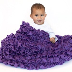 May include: A baby is sitting on a large, purple, ruffled blanket. The blanket is made of soft, plush fabric and has a lot of ruffles. The baby is wearing a white shirt and has a happy expression on their face.