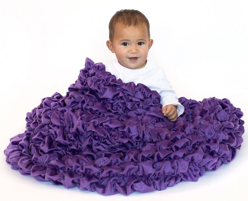 May include: A baby is sitting on a large, purple, ruffled blanket. The blanket is made of soft, plush fabric and has a lot of ruffles. The baby is wearing a white shirt and has a happy expression on their face.