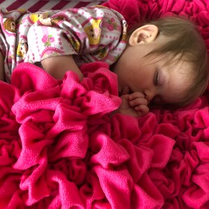 May include: A young child sleeping on a pink ruffled blanket. The child is wearing a pink and white patterned onesie.