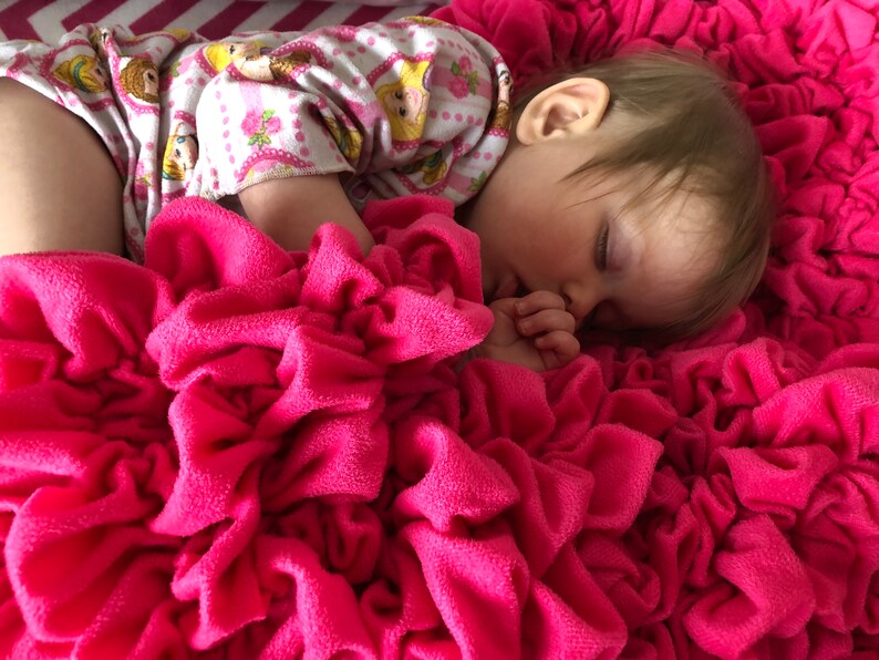 May include: A young child sleeping on a pink ruffled blanket. The child is wearing a pink and white patterned onesie.