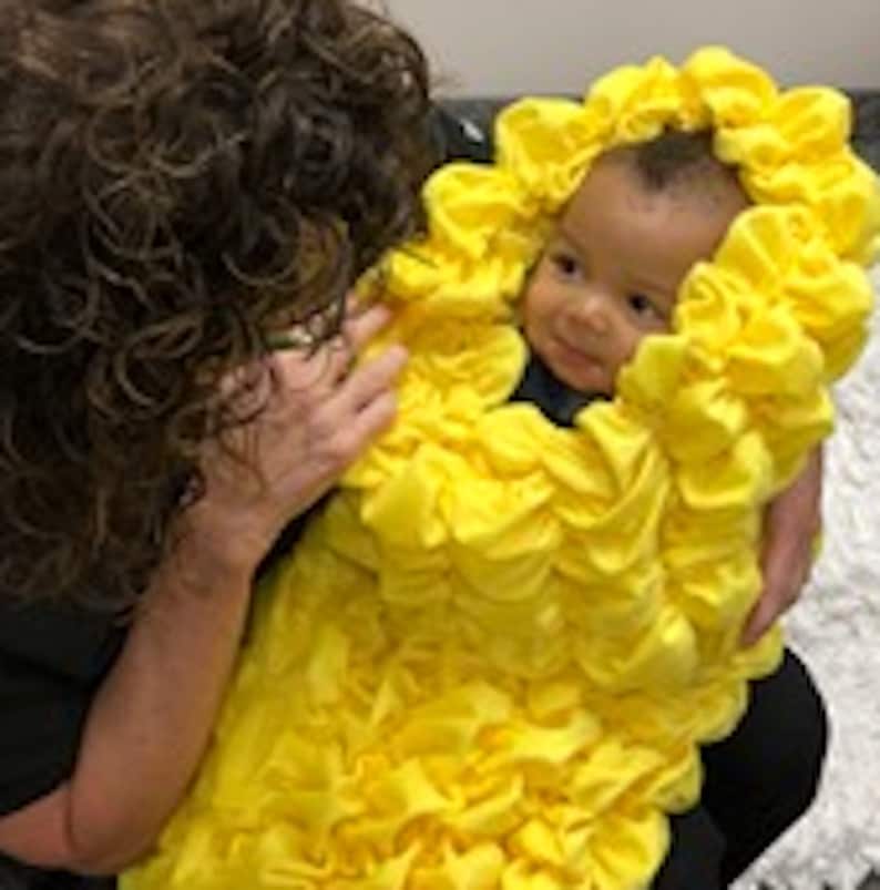 May include: A baby wearing a bright yellow ruffled costume resembling a flower. The baby is being held by an adult with dark curly hair.