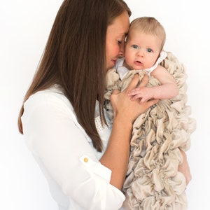May include: A woman gently cradles a baby wrapped in a beige ruffled blanket. The baby is looking up at the woman with big blue eyes.