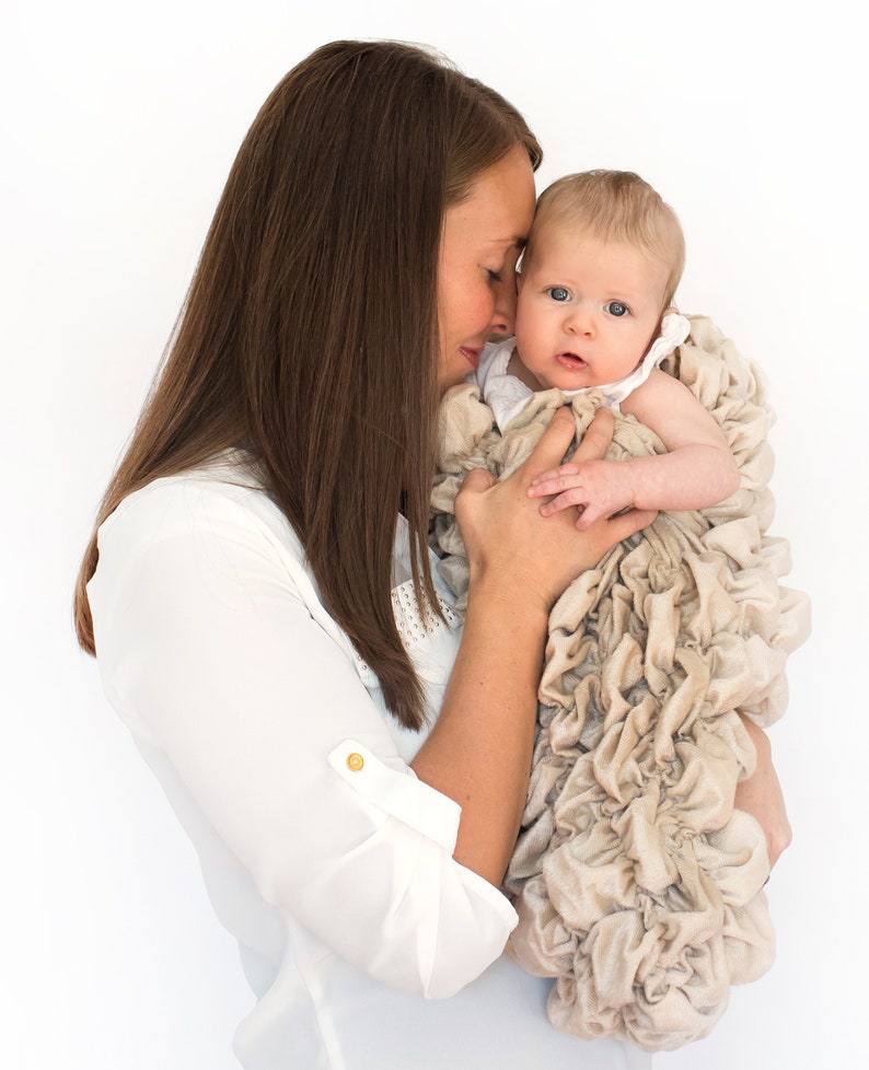 May include: A woman gently cradles a baby wrapped in a beige ruffled blanket. The baby is looking up at the woman with big blue eyes.