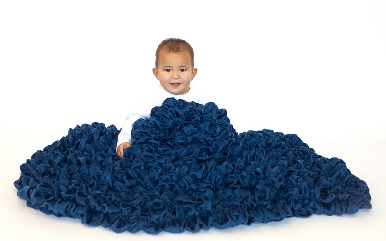 May include: A baby sitting on a large, blue, ruffled blanket. The blanket is made of soft, plush fabric and has a gathered, ruffled texture.