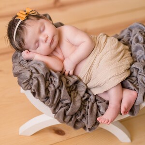 May include: A newborn baby sleeping peacefully in a brown and white swaddle, wrapped in a beige blanket, and lying on a gray, ruffled, fabric-covered prop. The baby is wearing a headband with a small, orange bow.