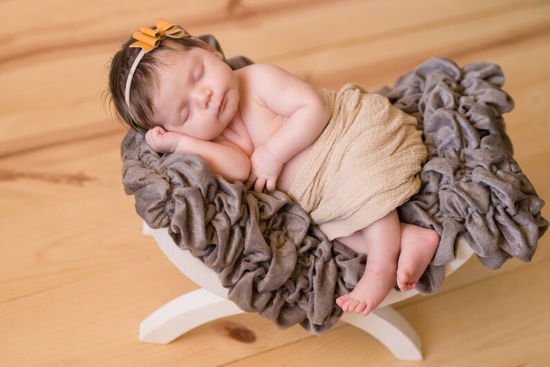 May include: A newborn baby sleeping peacefully in a brown and white swaddle, wrapped in a beige blanket, and lying on a gray, ruffled, fabric-covered prop. The baby is wearing a headband with a small, orange bow.