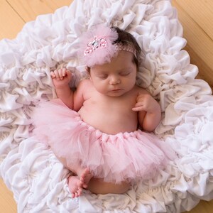 May include: A sleeping baby girl wearing a pink tutu and a sparkly headband with a white flower on a white ruffled surface.