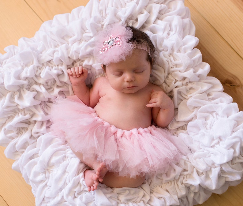 May include: A sleeping baby girl wearing a pink tutu and a sparkly headband with a white flower on a white ruffled surface.