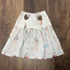 May include: White and patterned skirt with a dog print. The skirt has a white top with two brown circular accents. The bottom of the skirt is a light color with a repeating pattern of dogs in various colors.