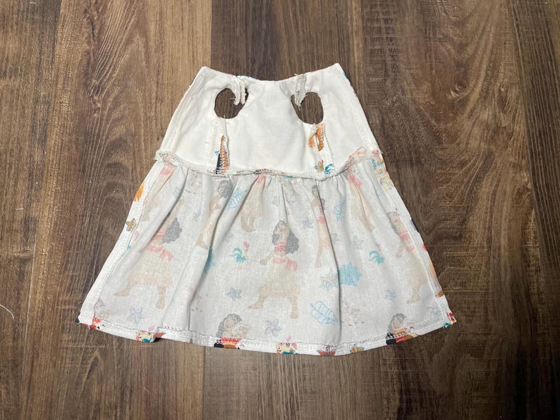 May include: White and patterned skirt with a dog print. The skirt has a white top with two brown circular accents. The bottom of the skirt is a light color with a repeating pattern of dogs in various colors.