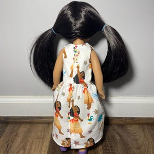 May include: A doll wearing a white dress with a colorful print. The dress features illustrations of a woman in a traditional outfit, a rooster, and other tropical elements. The doll has long black hair styled in two ponytails and is wearing purple shoes.