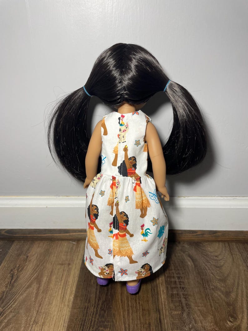 May include: A doll wearing a white dress with a colorful print. The dress features illustrations of a woman in a traditional outfit, a rooster, and other tropical elements. The doll has long black hair styled in two ponytails and is wearing purple shoes.