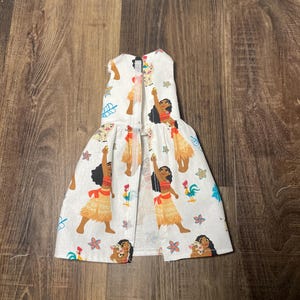 May include: White pet dress featuring a repeating pattern of a cartoon woman in a red top and yellow skirt, with a pig, rooster, and floral accents. The dress has a sleeveless design and a flared skirt.