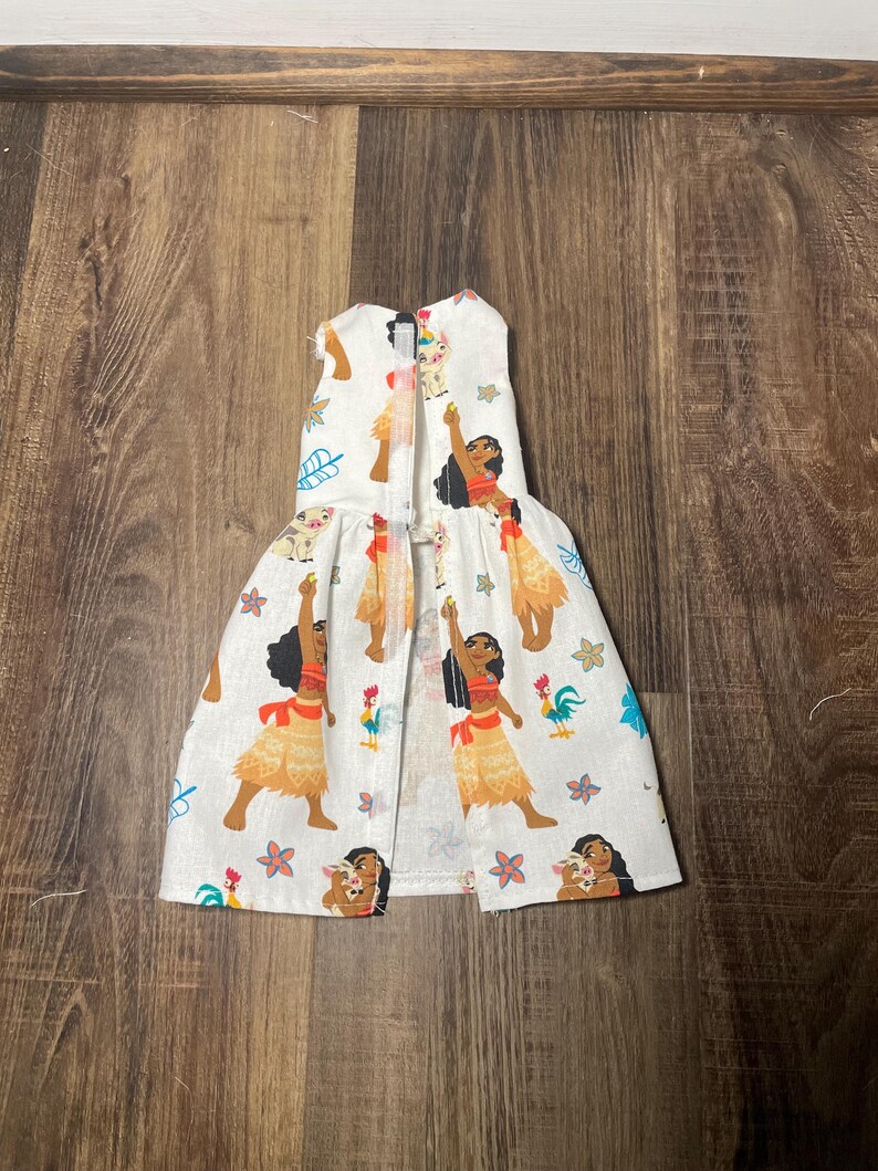 May include: White pet dress featuring a repeating pattern of a cartoon woman in a red top and yellow skirt, with a pig, rooster, and floral accents. The dress has a sleeveless design and a flared skirt.