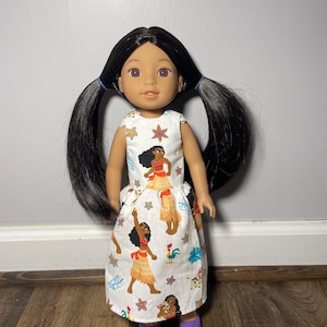 May include: A doll with black hair in pigtails wearing a white dress with a colorful print. The dress features images of a cartoon character in a yellow skirt and red top. The doll is wearing purple shoes.