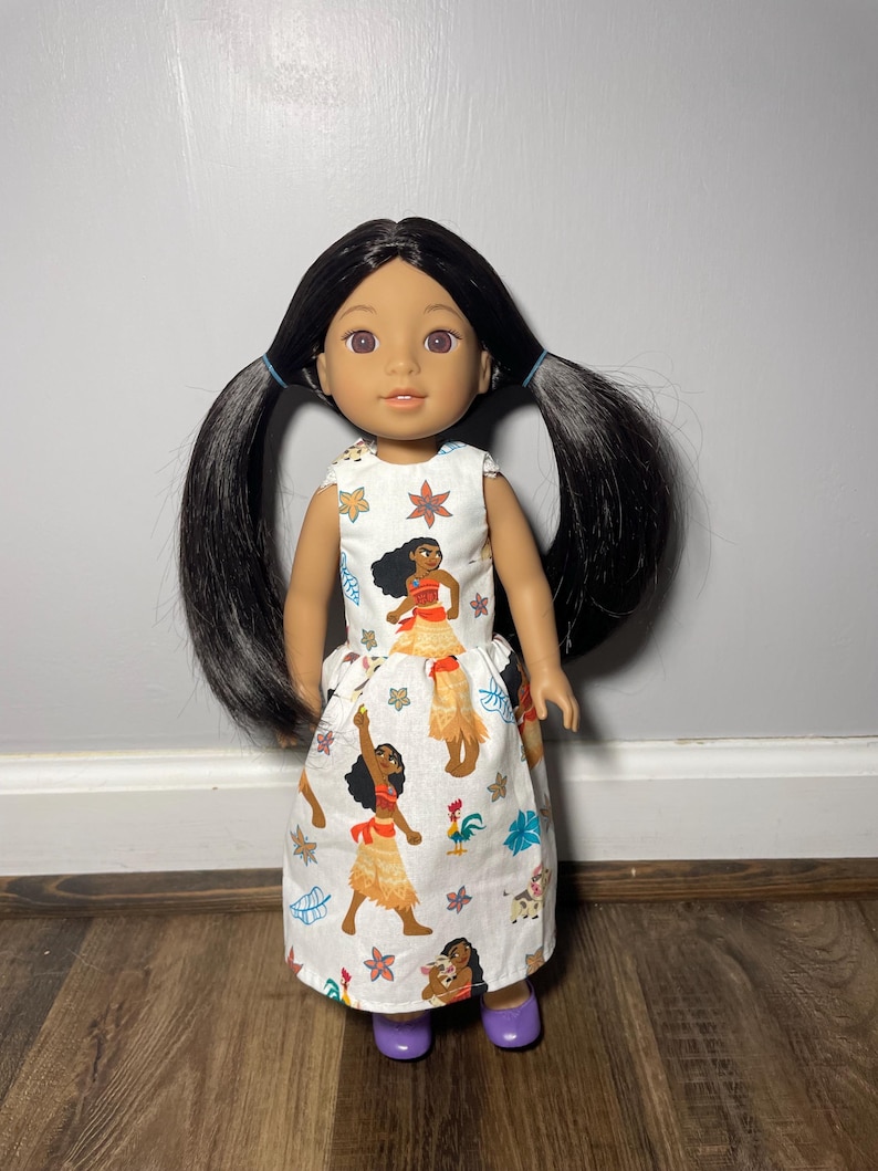 May include: A doll with black hair in pigtails wearing a white dress with a colorful print. The dress features images of a cartoon character in a yellow skirt and red top. The doll is wearing purple shoes.