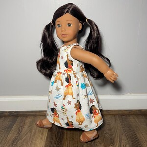 May include: A doll with dark hair in pigtails wearing a white dress with a colorful print of a woman in a grass skirt. The doll is also wearing brown sandals. The doll is standing on a wooden floor.