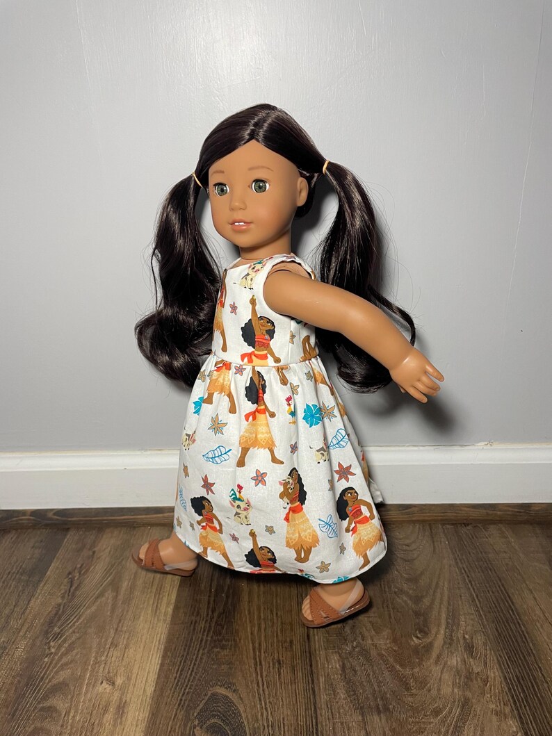 May include: A doll with dark hair in pigtails wearing a white dress with a colorful print of a woman in a grass skirt. The doll is also wearing brown sandals. The doll is standing on a wooden floor.