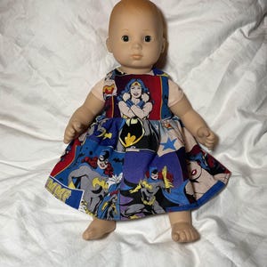 May include: A doll wearing a colorful dress featuring comic book characters such as Wonder Woman and Batgirl. The dress has a blue background with red, yellow, and white accents. The doll has blonde hair and is on a white background.