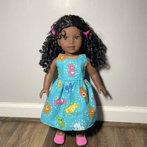May include: A doll with dark skin and curly black hair wearing a light blue dress with a colorful cartoon bear pattern. The doll has pink shoes and pink hair clips. The dress has a fitted bodice and a flared skirt.