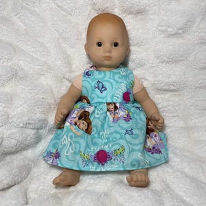 May include: A baby doll with a light skin tone wearing a sleeveless dress made of light blue fabric. The dress features a repeating pattern of a cartoon character, purple butterflies, and pink amulet icons, with the text Amulet printed on the fabric.