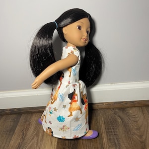 May include: A doll with black hair in pigtails wearing a white dress with a colorful print of characters. The doll has purple shoes. The dress features a pattern of cartoon characters, flowers, and leaves.