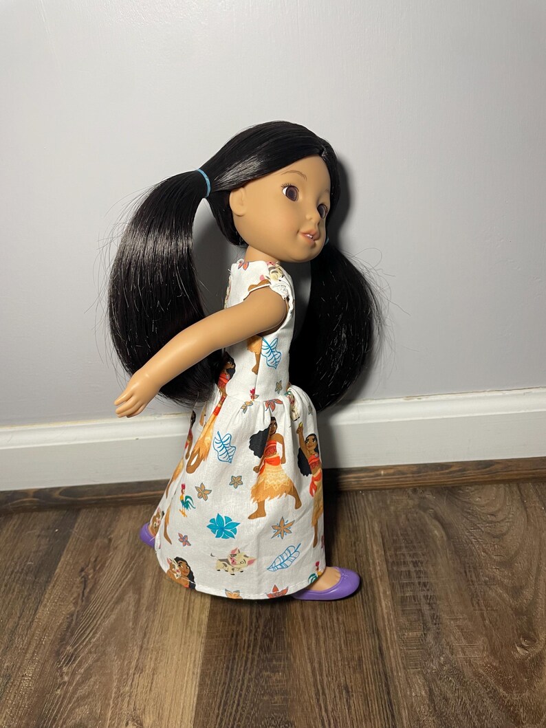 May include: A doll with black hair in pigtails wearing a white dress with a colorful print of characters. The doll has purple shoes. The dress features a pattern of cartoon characters, flowers, and leaves.