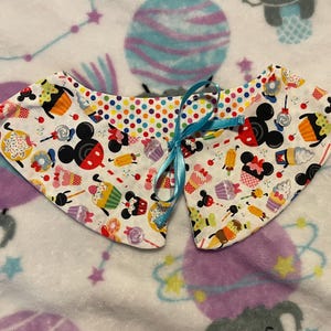 Puede incluir: Un cuello de tela blanco con un estampado colorido de personajes de Mickey Mouse, cupcakes y otros dulces. El cuello tiene un lazo azul para atar.