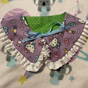May include: A lavender collar with a Hello Kitty print, green polka dot lining, and white ruffled trim. A light blue ribbon is tied at the center. The collar is designed for pets.