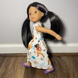 May include: A doll with long black hair in pigtails, wearing a white dress with a colorful print featuring cartoon characters and purple shoes. The doll has brown eyes and is posed as if walking.