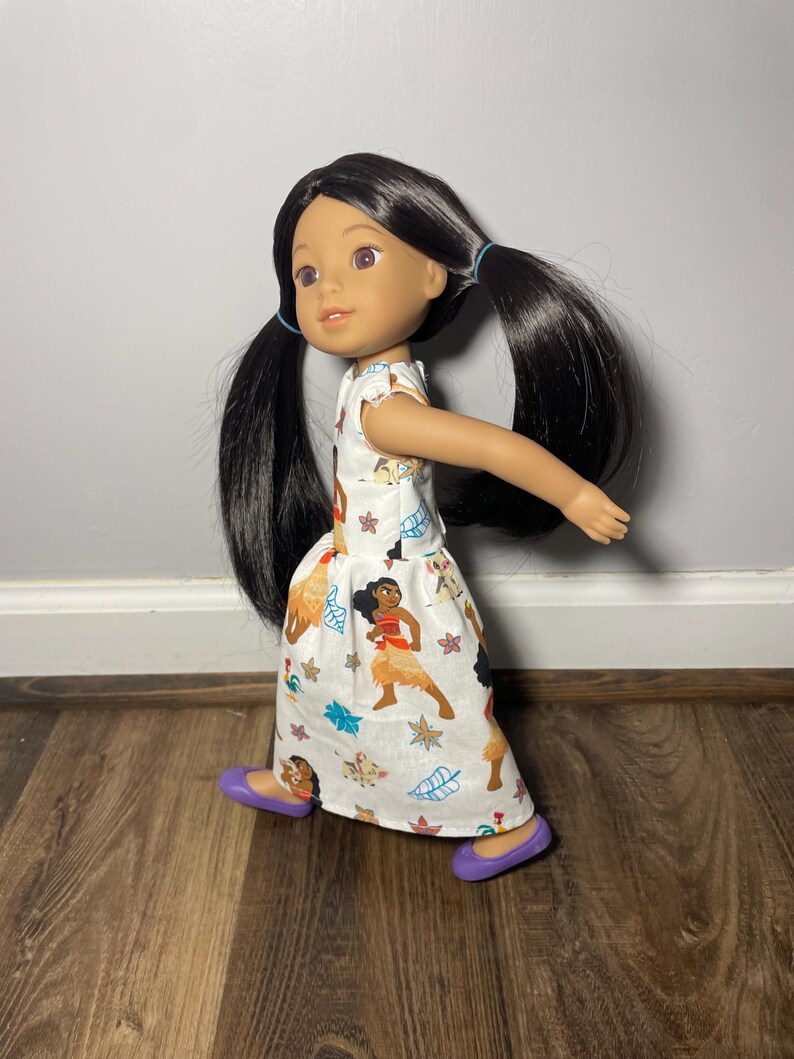 May include: A doll with long black hair in pigtails, wearing a white dress with a colorful print featuring cartoon characters and purple shoes. The doll has brown eyes and is posed as if walking.