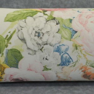 May include: A white cylindrical pillow with a floral pattern featuring pink, yellow, and blue flowers. The pillow has a green and gold trim.