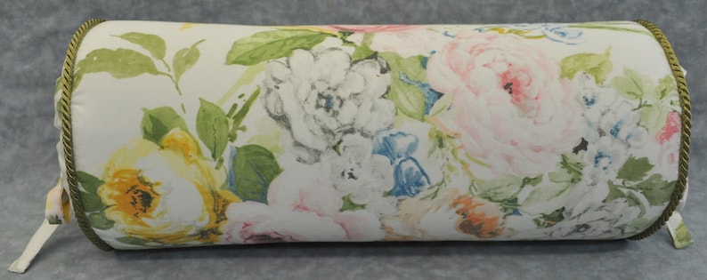 May include: A white cylindrical pillow with a floral pattern featuring pink, yellow, and blue flowers. The pillow has a green and gold trim.