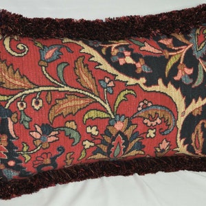 Puede incluir: Coj&iacute;n decorativo rectangular con fondo rojo y estampado floral en tonos azules, verdes y beige. El coj&iacute;n tiene un borde de flecos rojo oscuro.